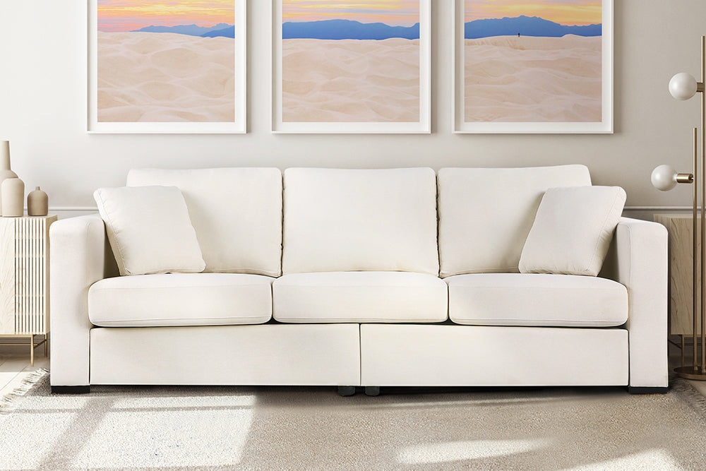 In the living room, there is a straight front view of a modern, beige, three-seat, eva modern fabric sofa with two side cushions.