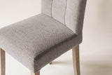 Close-up of one light grey woven fabric Adelina dining chair on an off-white backdrop: the box seat with its edge seam, the stitched panels of the back and the tops of the pale oak legs