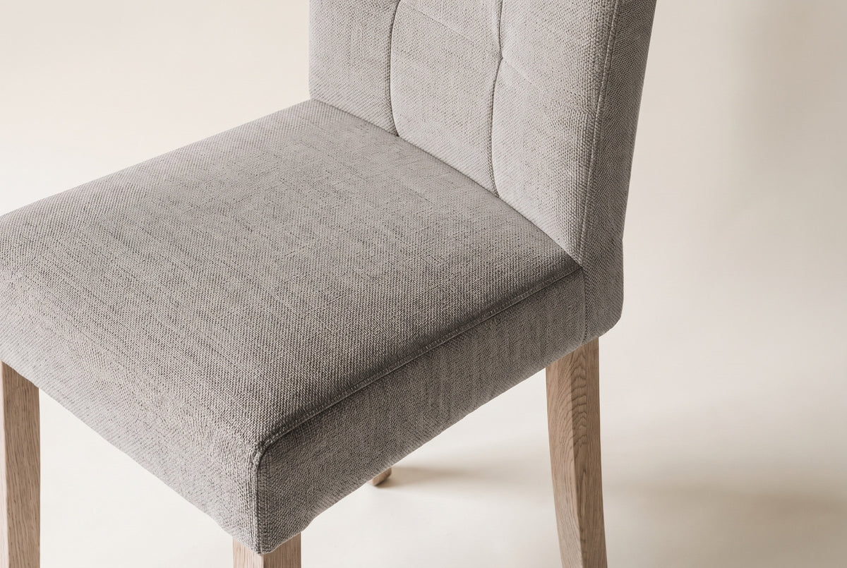 Close-up of one light grey woven fabric Adelina dining chair on an off-white backdrop: the box seat with its edge seam, the stitched panels of the back and the tops of the pale oak legs