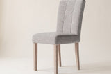 Side view of one light grey Adelina upholstered dining chair on an off-white backdrop, with the tall stitched back leaning back, the box seat and four square pale oak legs