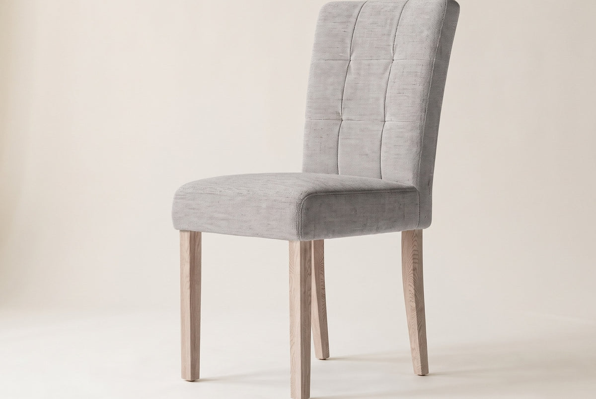 Side view of one light grey Adelina upholstered dining chair on an off-white backdrop, with the tall stitched back leaning back, the box seat and four square pale oak legs