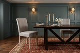 Two light grey Adelina upholstered dining chairs with pale oak legs at a long dark wood trestle table with three lit candles in pewter candlesticks, two place settings and white sweet peas, in a teal-green panelled dining room with brass wall lights and a red patterned rug in the evening