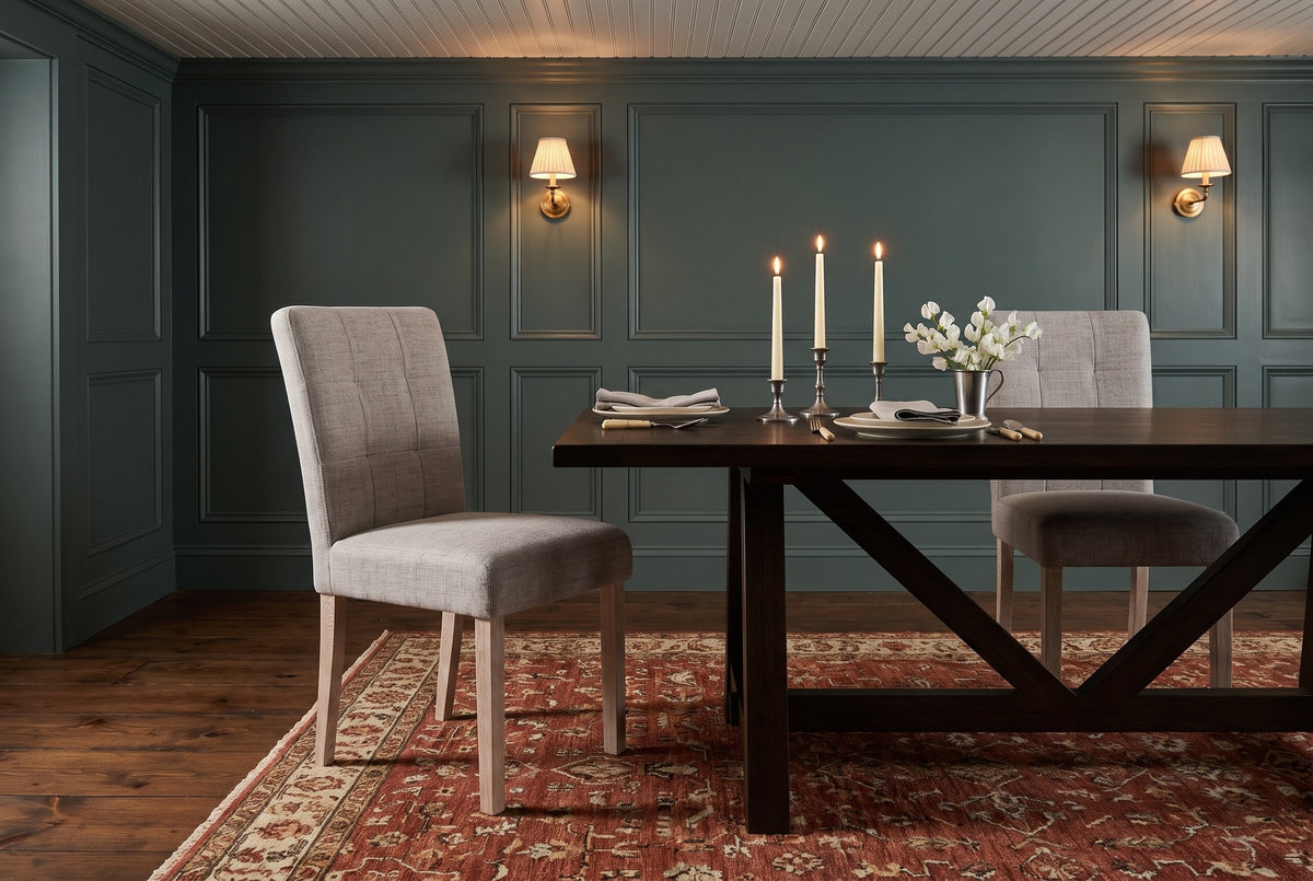 Two light grey Adelina upholstered dining chairs with pale oak legs at a long dark wood trestle table with three lit candles in pewter candlesticks, two place settings and white sweet peas, in a teal-green panelled dining room with brass wall lights and a red patterned rug in the evening