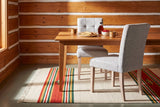 Two light grey Adelina upholstered dining chairs with pale oak legs at a honey-toned wood table with crepes, a bottle of maple syrup, a bowl of raspberries and two caramel mugs, in a log-walled breakfast room with a sage-grey painted floor and a striped wool rug in morning sun