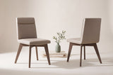 Two Aileen dining chairs in beige leather, one seen from the front and one from behind, with an olive sprig in a sage vase on travertine blocks between them