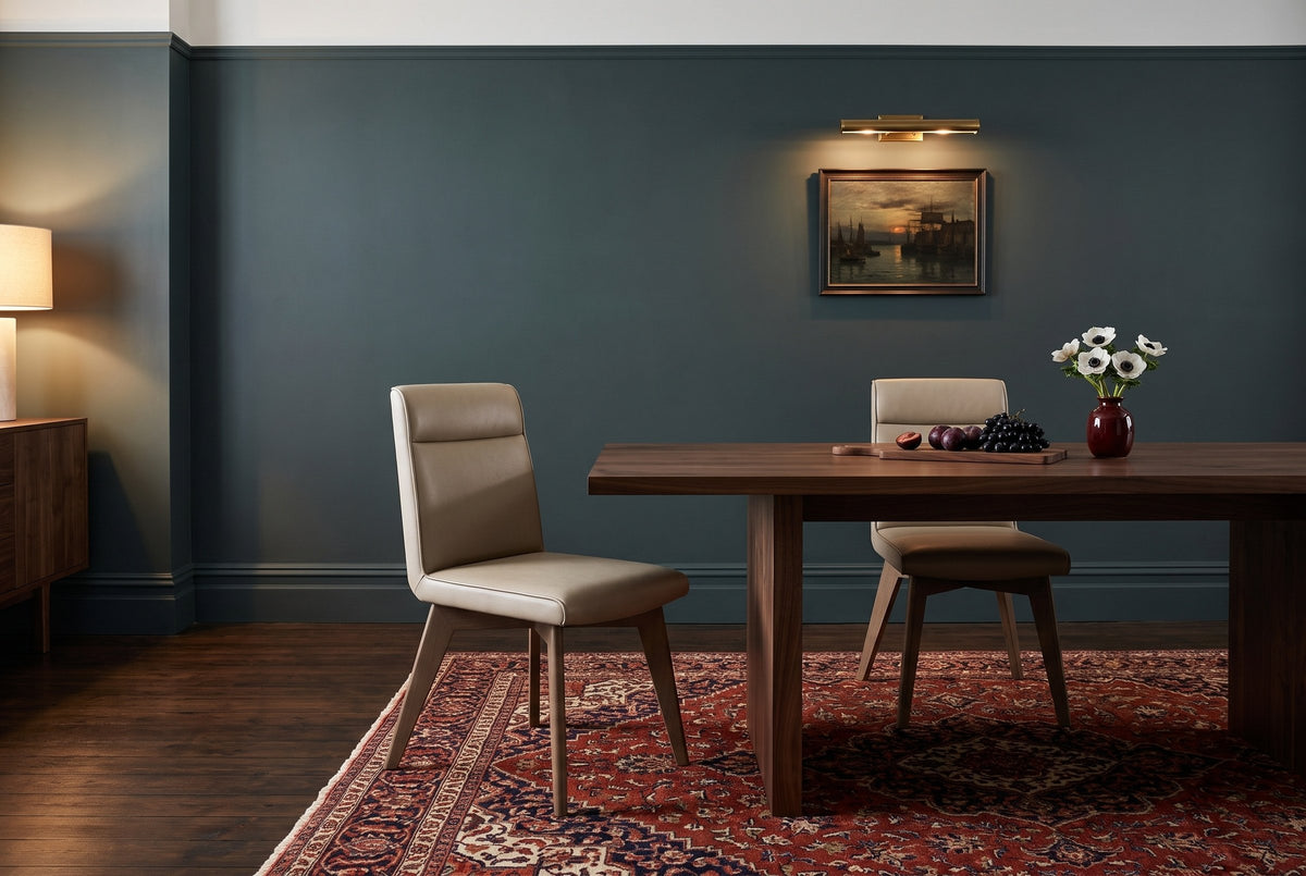 Two beige Aileen leather dining chairs with ash legs at a long walnut table with a walnut board of plums and black grapes and a dark red vase of white anemones, in a deep teal dining room with a brass picture light over a harbour painting, a lit lamp on a walnut sideboard and a red patterned rug on a dark wood floor in the evening