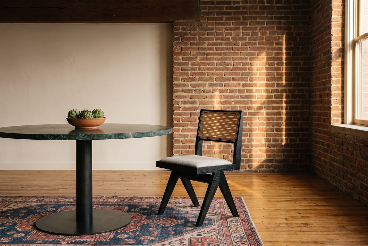 Alaya black oak dining chair with a cane back and oatmeal seat beside a round green marble table with a bowl of artichokes, by an exposed brick wall in golden-hour light