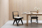 Two Alaya black oak dining chairs with cane backs and oatmeal seats at a white oak table with white bowls, a linen napkin and white cosmos, in a pale oak-panelled room with a limestone floor