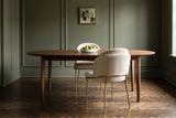 Alexander ivory fabric dining chair with brass legs seen from behind at an oval walnut table with a bowl of pears, a second chair across, in an olive-green panelled dining room with a herringbone floor