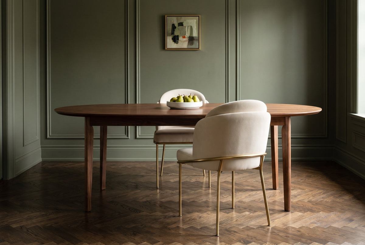Alexander ivory fabric dining chair with brass legs seen from behind at an oval walnut table with a bowl of pears, a second chair across, in an olive-green panelled dining room with a herringbone floor