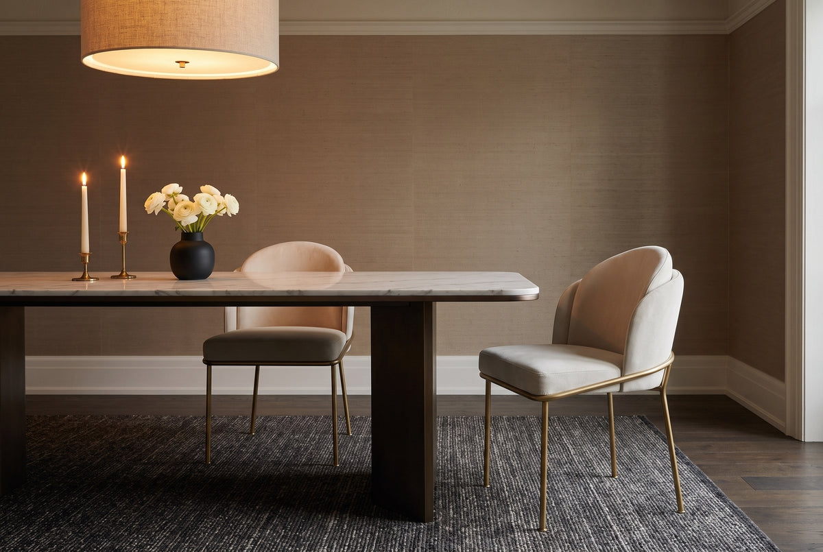 Two Alexander ivory fabric dining chairs with brass legs at a white marble table on a dark base with candles, white ranunculus and a linen drum pendant, taupe grasscloth walls and a charcoal rug, in the evening
