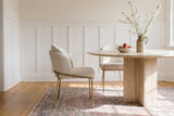 Two Alexander ivory fabric dining chairs with brass legs at a round travertine pedestal table with a bowl of peaches and a blossom branch in a stoneware jug, white wainscoting and a rose and blue rug, in morning light