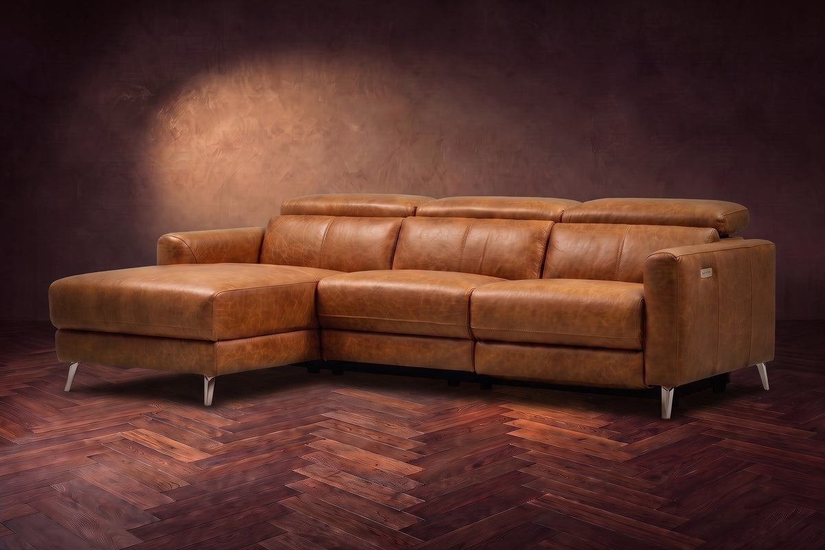 The Andria distressed Italian leather power reclining sectional on a walnut herringbone floor against a brown plaster wall, three-quarter view, headrests down and the footrest closed. Shown in Antiqued Caramel, Left chaise.