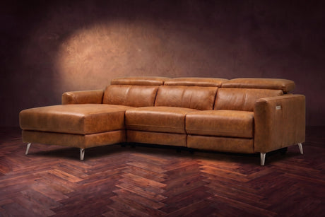 The Andria distressed Italian leather power reclining sectional on a walnut herringbone floor against a brown plaster wall, three-quarter view, headrests down and the footrest closed. Shown in Antiqued Caramel, Left chaise.