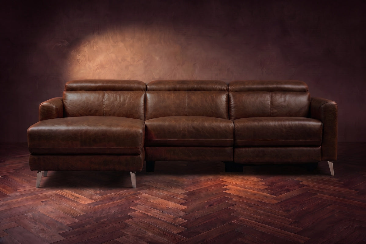The Andria distressed leather reclining sectional seen from the front on a walnut herringbone floor, headrests down and the footrest closed. Shown in Burnished Chestnut, Left chaise.