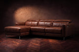 The Andria distressed Italian leather power reclining sectional on a walnut herringbone floor against a brown plaster wall, three-quarter view, headrests down and the footrest closed. Shown in Burnished Chestnut, Left chaise.