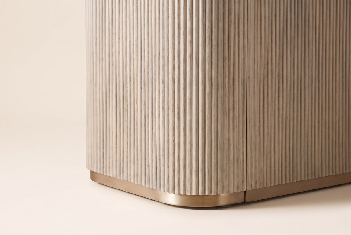 Close-up of the Aquileia pedestal's lower corner in Natural: vertical flutes wrapping the rounded corner above a brass-tone m