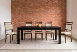 Five Arden dining chairs around a long black table on a wool rug in front of a red-brick wall, with a bowl of oranges