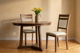 Two Arden dining chairs at a round walnut table with yellow tulips, one pulled out, against an ochre limewashed wall on an oak parquet floor
