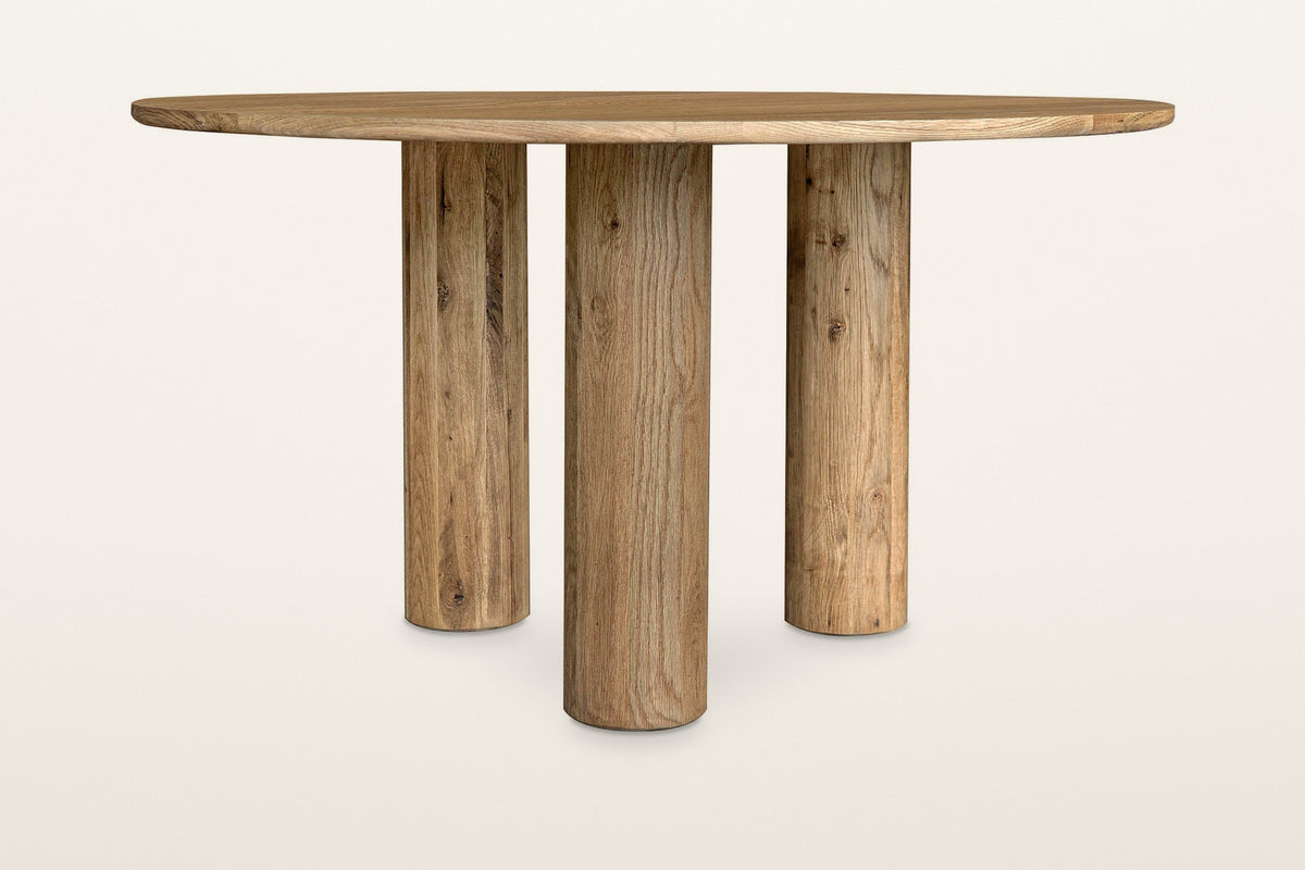 Atzara round dining table in Natural, level side view on a warm white studio backdrop: three column legs set well in under th