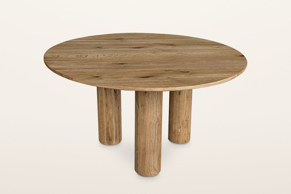 Atzara round oak dining table in Natural, three-quarter view on a warm white studio backdrop: a thin 55 in round top on three