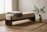 The Bassano bench in beige loop-yarn boucle on dark rubberwood slab legs with a folded wool throw, against a whitewashed brick wall on old oak floorboards and a jute rug in an Amsterdam canal house