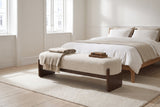 The Bassano bench in beige loop-yarn boucle on dark rubberwood slab legs at the foot of a low oak bed with white and oat linen, in a bright Copenhagen apartment bedroom with pale plank floors and a wool rug