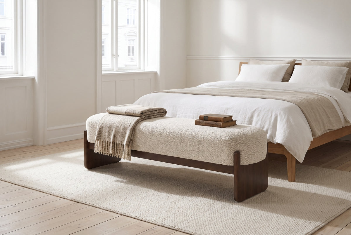 The Bassano bench in beige loop-yarn boucle on dark rubberwood slab legs at the foot of a low oak bed with white and oat linen, in a bright Copenhagen apartment bedroom with pale plank floors and a wool rug