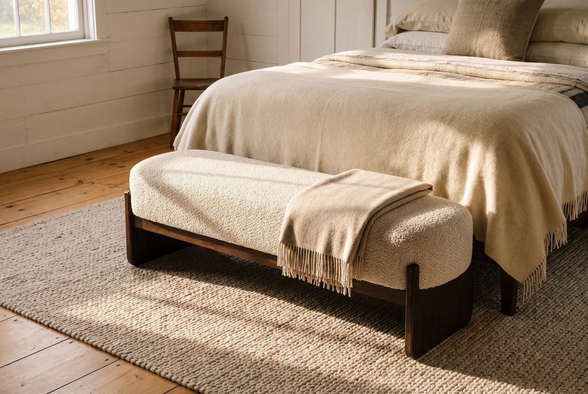 The Bassano bench in beige loop-yarn boucle on dark rubberwood slab legs at the foot of a bed with a cream wool blanket, in a Vermont farmhouse bedroom with board walls and wide pine floors
