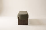 The dark green Bassano bench seen from the end: the rounded boucle seat on one dark rubberwood slab leg