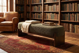 The Bassano bench in dark green loop-yarn boucle on dark rubberwood slab legs in front of walnut bookshelves on a red antique rug, beside a tan leather armchair, in a Hudson Valley library