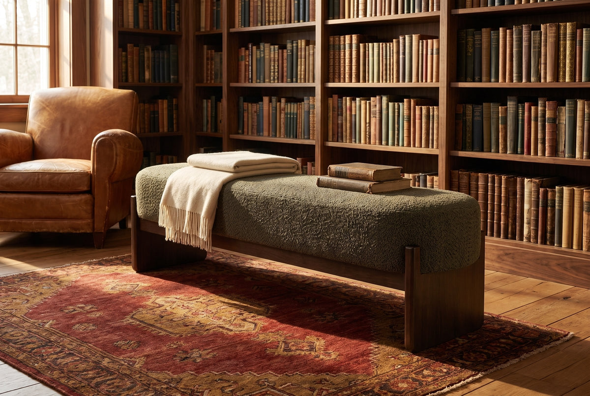 The Bassano bench in dark green loop-yarn boucle on dark rubberwood slab legs in front of walnut bookshelves on a red antique rug, beside a tan leather armchair, in a Hudson Valley library