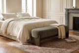 The Bassano bench in dark green loop-yarn boucle on dark rubberwood slab legs at the foot of a bed in ivory linen, in a Paris apartment bedroom with panelled walls, herringbone parquet and a grey marble fireplace