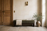 The Bassano bench in dark green loop-yarn boucle on dark rubberwood slab legs along a pale limestone wall on a worn stone floor, beside an old oak door, in a Paris entrance hall