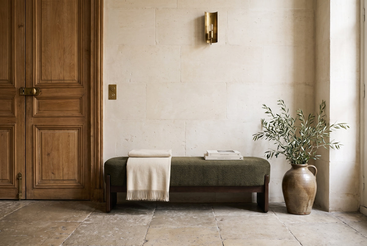 The Bassano bench in dark green loop-yarn boucle on dark rubberwood slab legs along a pale limestone wall on a worn stone floor, beside an old oak door, in a Paris entrance hall