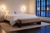 Bassano bench at the foot of a bed in a Montreal loft bedroom at dusk, white brick wall and two wall lights