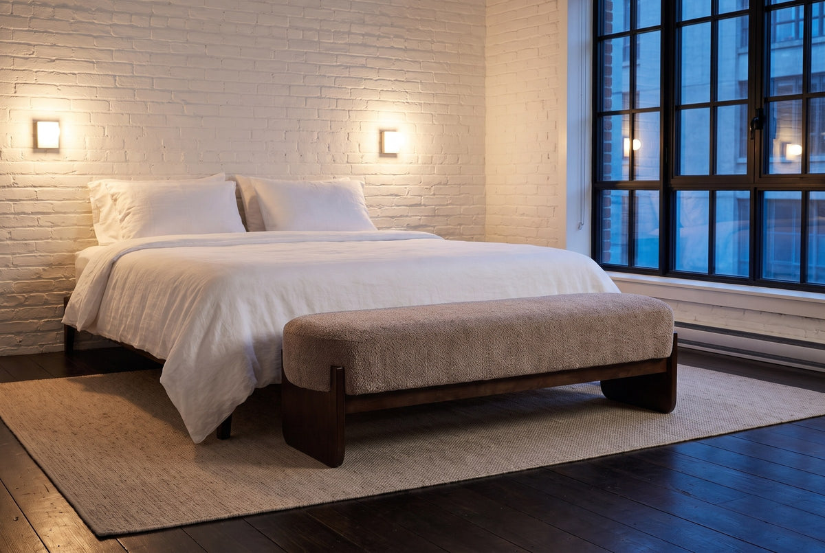 Bassano bench at the foot of a bed in a Montreal loft bedroom at dusk, white brick wall and two wall lights