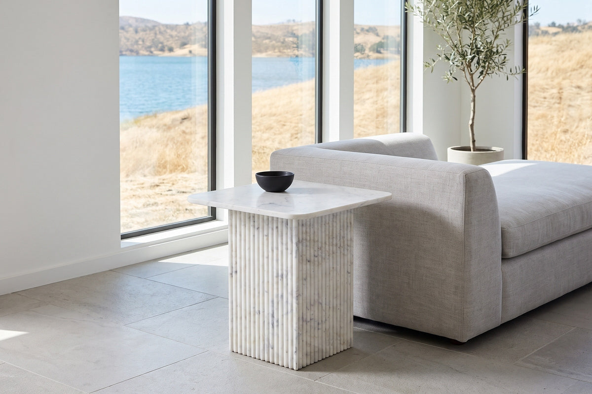 Camerota white marble side table at the end of a grey linen sofa in a Kelowna lake house, a black stoneware bowl on top