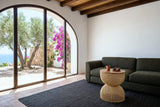 The Canale side table in Natural, seen from a high corner, beside an olive-green linen sofa in a Mojacar courtyard house