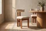 Two Carlo dining chairs in oak and ivory boucle, one on an oat wool rug and one behind a round white oak pedestal table with a stoneware vase of olive branches and a ribbed glass, against a warm plaster wall in late-afternoon window light