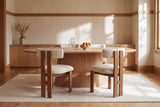 Four Carlo dining chairs in oak and ivory boucle tucked in around an oval white oak pedestal table with a glass carafe and a bowl of clementines, on a cream wool rug, with dried grasses in a stoneware jug on a long oak sideboard, oak trim and sunlit plaster walls