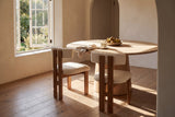 Two Carlo dining chairs in wire-brushed oak and ivory boucle at a round white oak pedestal table with a bowl of pears, a cup and saucer and a linen napkin, in a sunlit plaster room with an open casement window onto a garden, an arched doorway and a wide-plank oak floor