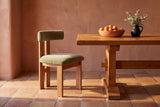 A Carlo oak dining chair with a sage-olive velvet bolster back and seat beside a plank-top trestle table on terracotta tiles, with clay plaster walls, a bowl of apricots and a black jug of sage in low warm sun