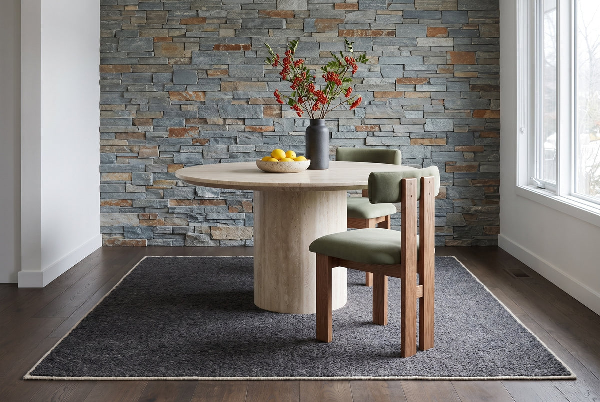 Two Carlo oak dining chairs with sage-olive velvet bolster backs and seats at a round travertine pedestal table on a charcoal rug, in front of a stacked stone wall, with a bowl of lemons and a vase of red berry branches