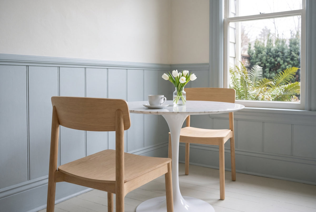 Two Casole dining chairs in natural oak at a small round marble table in a Victoria breakfast nook with pale blue-grey panell