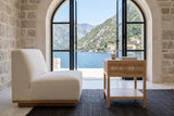Side view of the Cetara side table in Natural ash and wicker beside an ivory lounge chair in a stone house by the bay