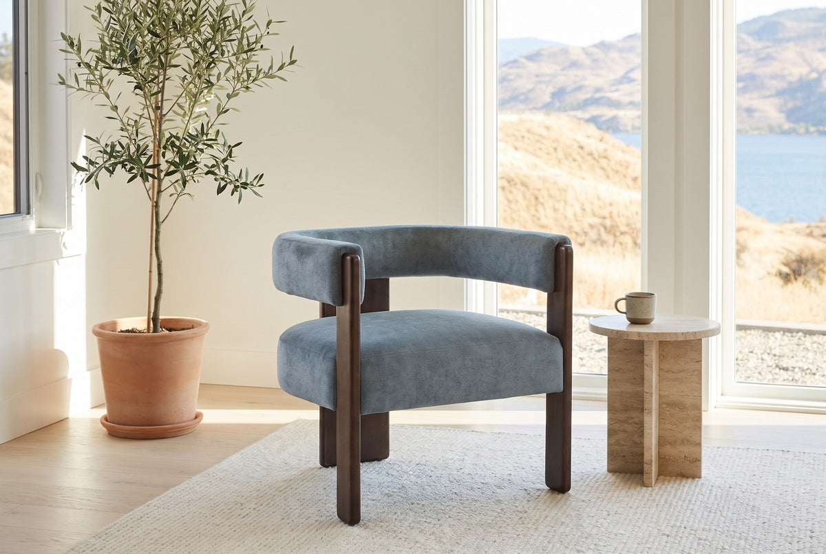 Chioggia accent chair in Blue by a lake-view window with a travertine side table and an olive tree