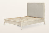 Chiusdino bed frame in whitewashed wood, three-quarter view on a warm white studio backdrop: a tall greige linen-look headboa