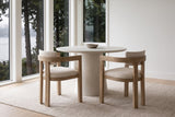 Two Cisternino chairs pulled out from a round plaster table beside tall windows onto cedar trees and an inlet