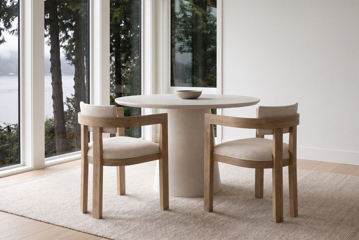 Two Cisternino chairs pulled out from a round plaster table beside tall windows onto cedar trees and an inlet